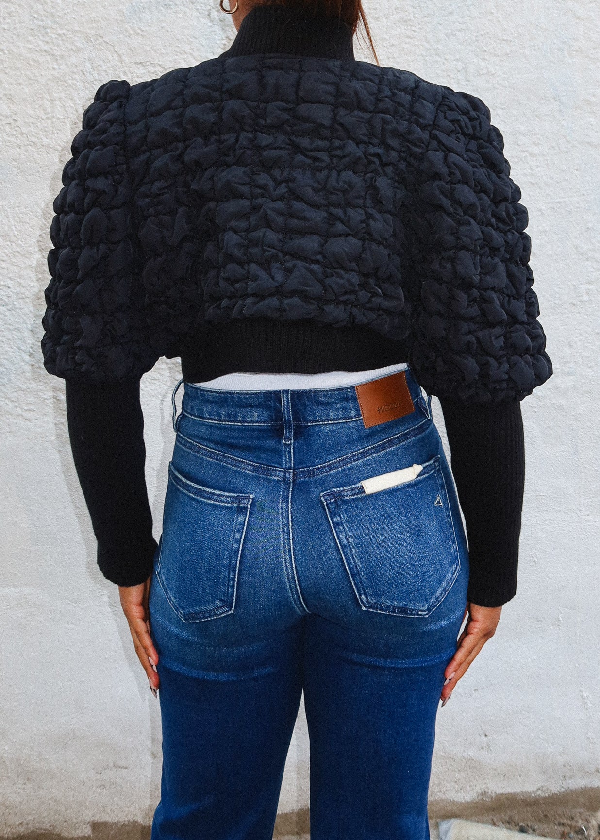 Person wearing a black textured jacket and blue jeans against a white wall